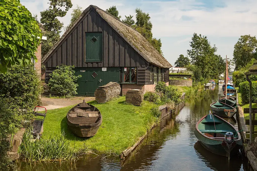Giethoorn'da kanal kıyısında kamış çatılı ahşap bir ev, çimenlikteki kayık ve kanalda sıralı tekneler